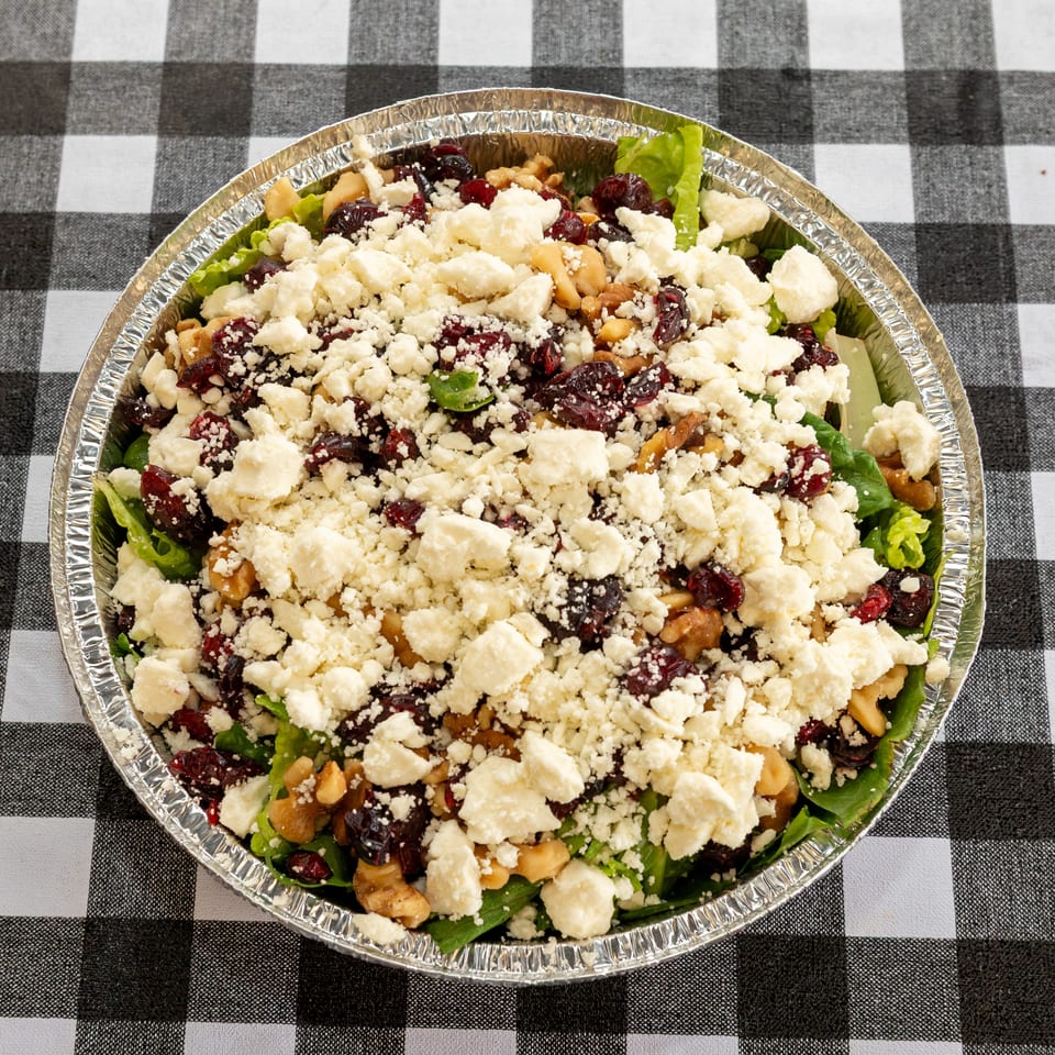 Salad with lettuce, cranberries, walnuts, and feta cheese in a foil container on a checkered tablecloth.