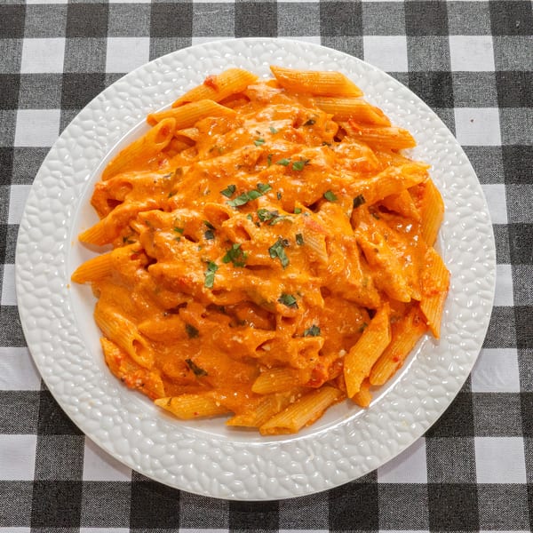 Penne pasta with creamy orange sauce and fresh herbs on a white plate over a checkered tablecloth.