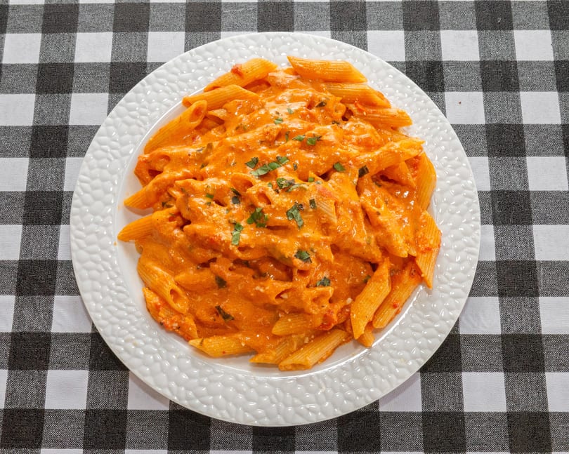 Penne pasta with creamy orange sauce and fresh herbs on a white plate over a checkered tablecloth.