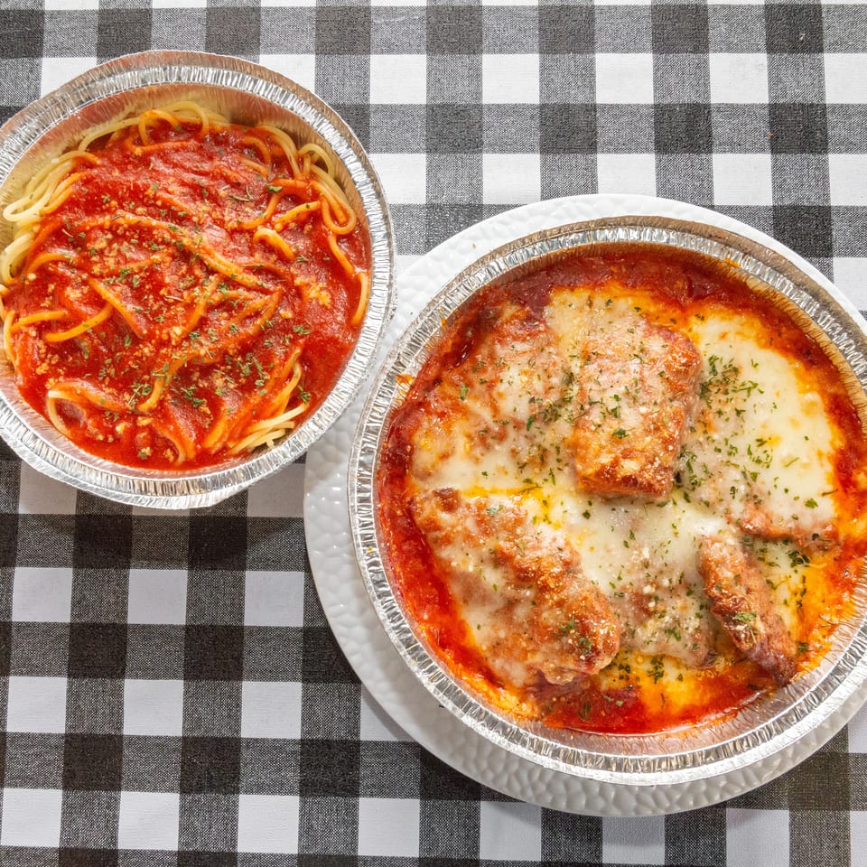 Two foil containers of Italian food, spaghetti with red sauce and chicken parmesan, on a checkered tablecloth.