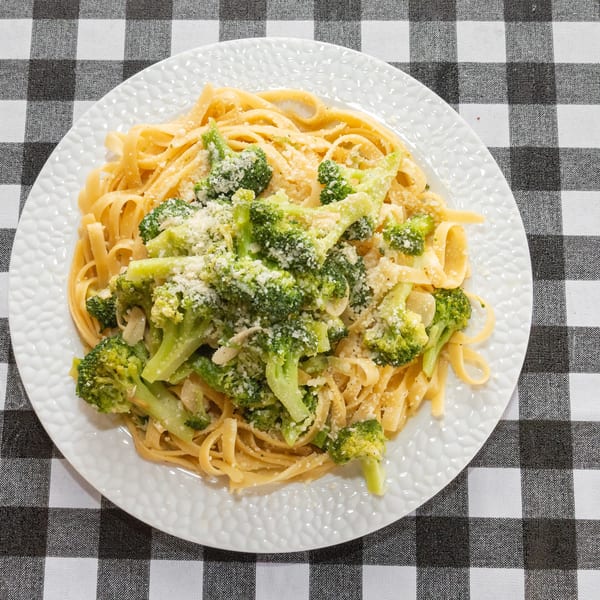 Fettuccine pasta with broccoli florets and grated cheese on a white plate, on a black and white checkered tablecloth.