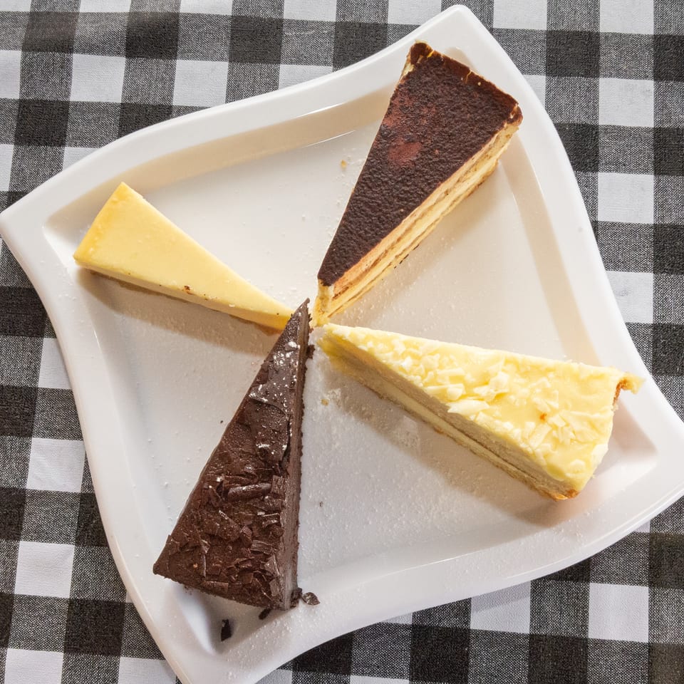 Four different dessert slices, including chocolate and cheesecake, arranged on a white plate over a checkered tablecloth.