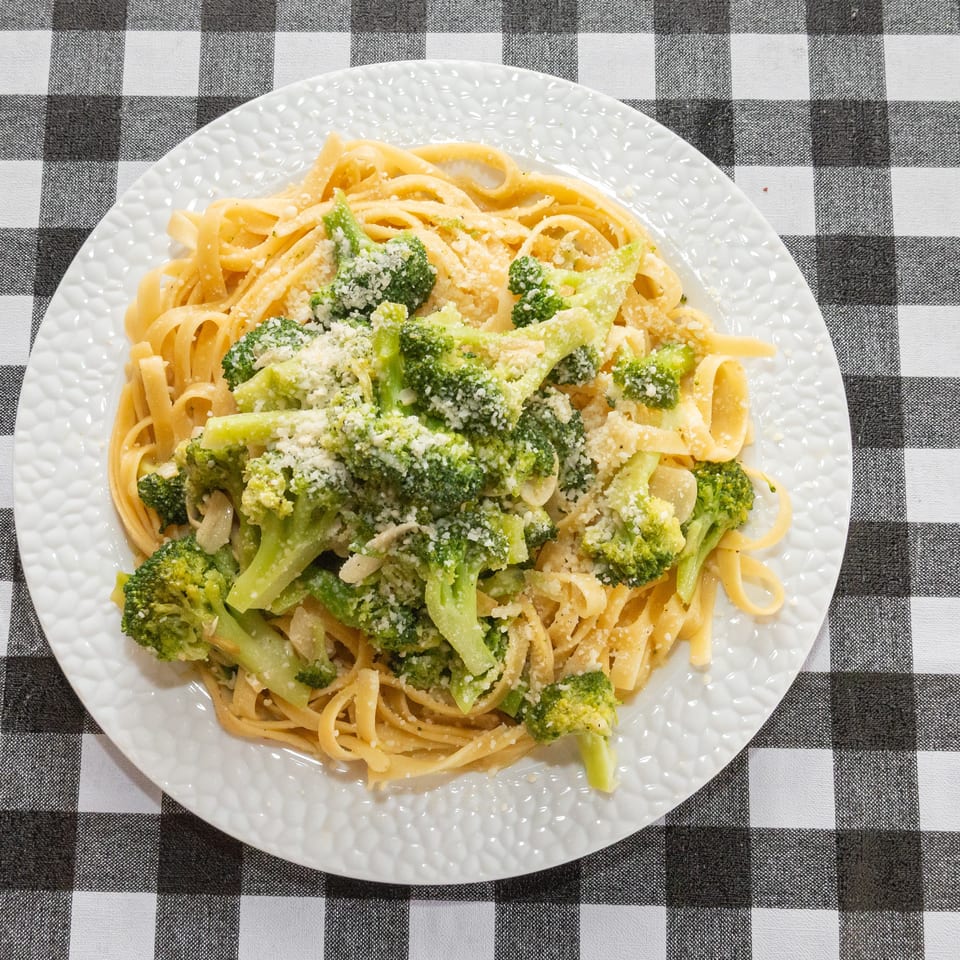 Fettuccine pasta with broccoli florets and grated Parmesan cheese on a white plate, on a checkered tablecloth.